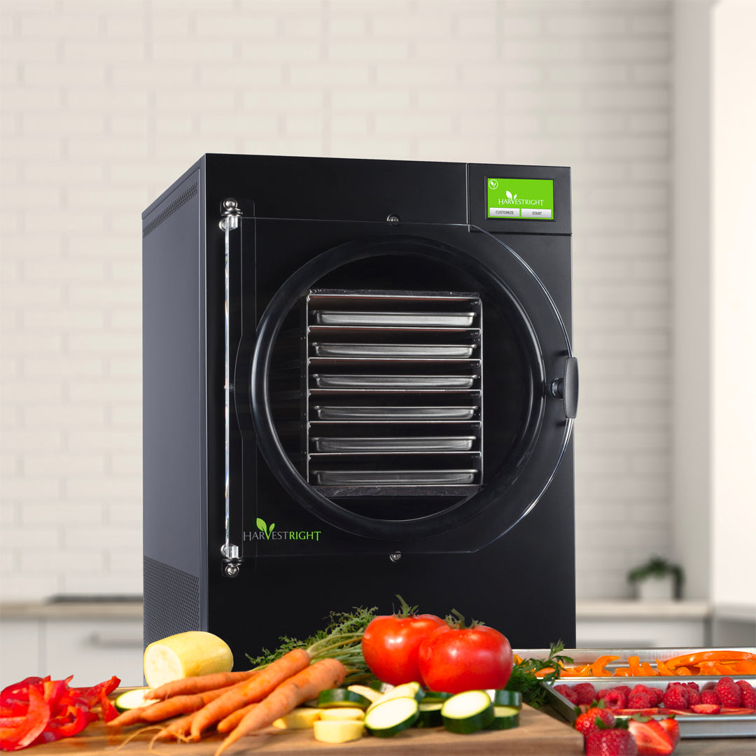 Black Harvest Right Large Pro home freeze dryer with six stainless steel trays visible through the round door, on a kitchen counter beside fresh garden produce